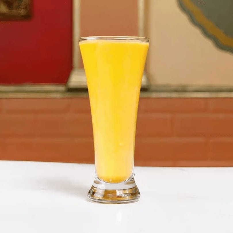 Tall glass filled with bright yellow mango lassi on a white table with a red and brown background.