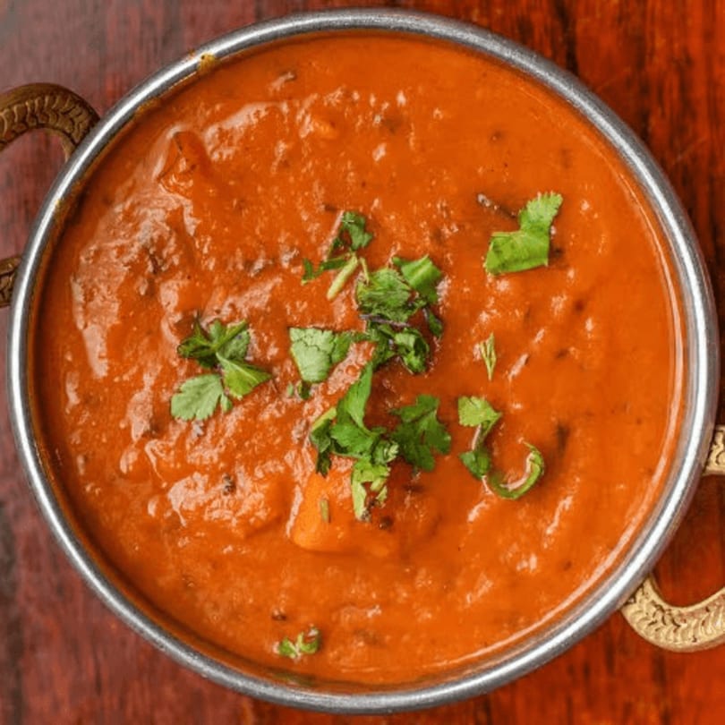 A metal bowl filled with vibrant orange curry, garnished with fresh green cilantro, on a wooden surface.